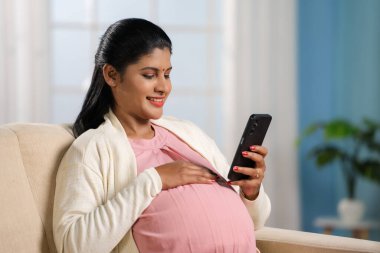 Mutlu Hintli hamile kadın evde cep telefonu kullanmakla meşgul - hamilelik uygulamaları, bebek hazırlıkları ve sosyal medya paylaşımı kavramı