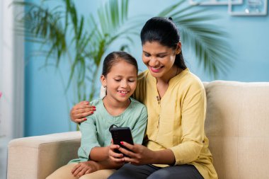 Mutlu Hintli anne evde koltukta otururken kızına cep telefonu gösteriyor. Teknoloji, rahatlama ve bağlanma kavramı.