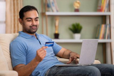 Mutlu gülümseyen Hintli adam evdeki koltukta dizüstü bilgisayarla alışveriş için online ödeme yapıyor. İnternet bankacılığı, ticaret ve güvenli online işlem kavramı..