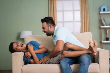 Neşeli Hintli baba evdeki kanepede oğluna sarılarak oynuyor. Aile bağı, zaman geçirme ve sevecenlik kavramı.