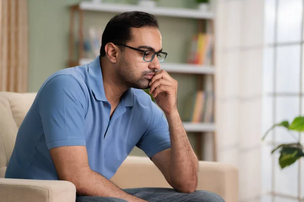 Worried indian man deep thinking while sitting on sofa at home - concept of financial problem, love breakup and mental illness