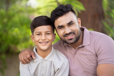 Mutlu gülümseyen Hintli baba ve oğlu birbirlerini kucaklamak için parka bakarlar. Aile birliği, kuşak ve babalık kavramı.