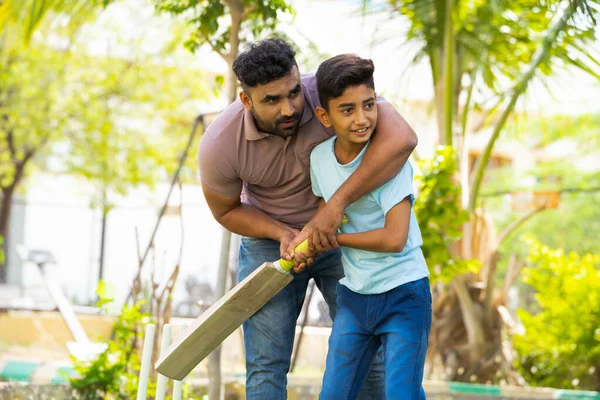 indian father teaching his son to playing cricket game at park - concept of Shared passion, family bonding and fatherhood.