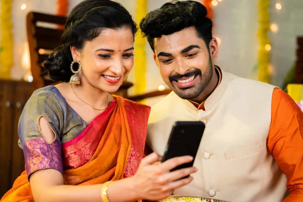 Happy young indian couple watching online shopping on mobile phone at home - concept of social media sharing, internet and sending festive wishes and greetings.