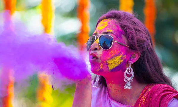 Young indian girl with sunglasses blowing holi colour powder from hand during on flower decoration background - concept of holi festival celebration, Indian tradition and colourful culture.