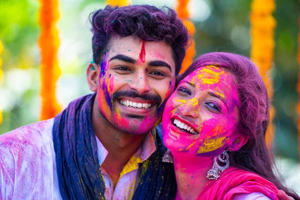Close up shot of happy young indian couple with holi colour on face by looking at camera during holi festival celebration - concept of freedom, togetherness and indian culture.