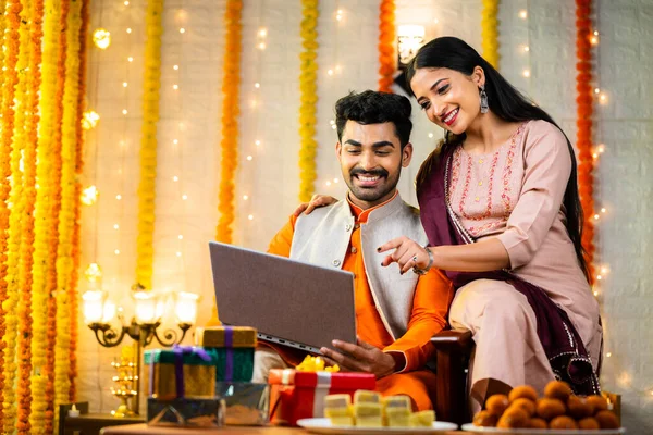 Happy young indian couple busy checking laptop during festival celebration at home - concept of sales or offers, buying new apartment and planning.
