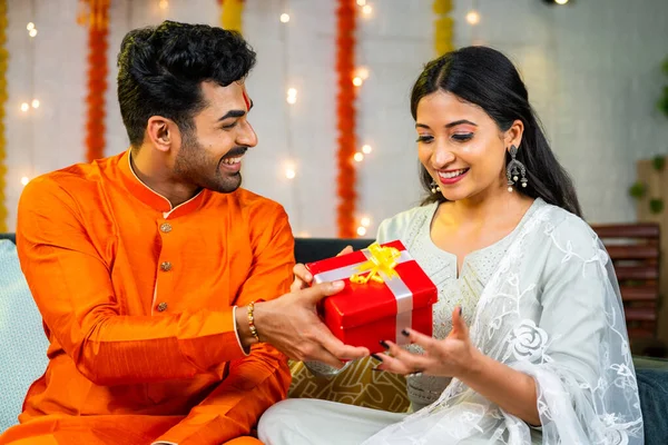 Happy young husband giving present to wife during festival celebration at home - brother giving gift to sister during raksha bandhan