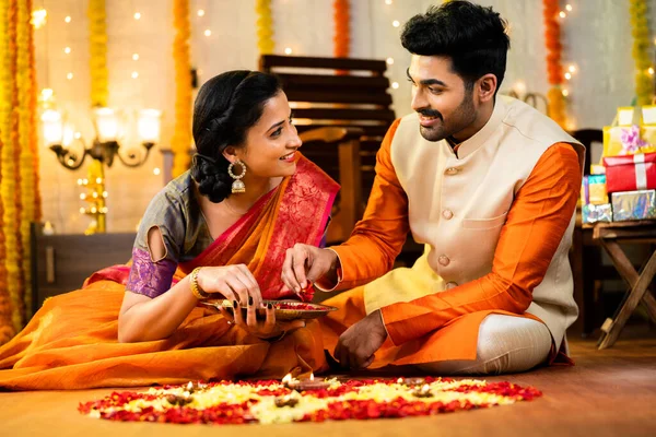 Happy indian couple in traditional ethnic wear decorating rangoli with flower for festival celebration on floor - concept of relationship, bonding, and religious ceremony