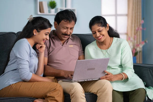 Happy indian middle aged parents with adult daughter using laptop on sofa at home - concept of Modern Family, Togetherness and Parental Support.