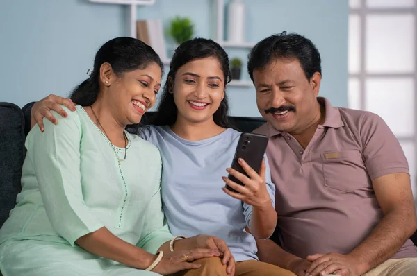 Happy indian daughter showing mobile phone to parents while sitting on sofa at home - concept of Parental Bonding, Technology and social media interaction.