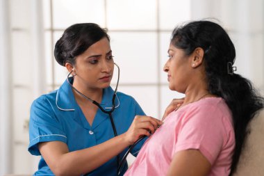 Hintli hemşire ya da doktor evdeki kanepede steteskop kullanan hasta kadını muayene ediyor. Tıbbi muayene, ev sağlığı ve sağlık değerlendirmesi kavramı..