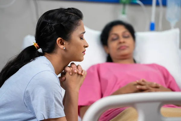 worried indian daughter praying for sick admitted mother for health recovery at hospital - concept of emotional hope, family togetherness and medical treatment.