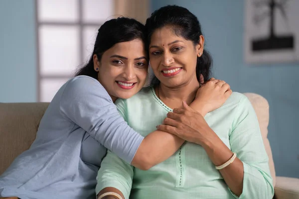 Happy indian mother hugging her daughter by looking camera while sitting on sofa at home - concept of Tender or Affection, Family Togetherness and Caring Connection