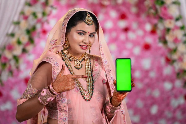 Indian miling young woman in Bridal wear on decorated background showing green screen mobile phone - concept of app promotion, advertisement and technology