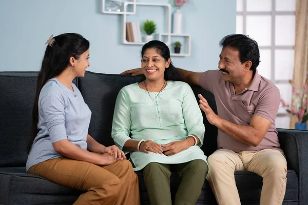 Happy smiling indian middle aged parents with daughter talking each other by looking at camera on sofa at home - concept of Parental Bonding, Family Conversation and Relaxed Discussion