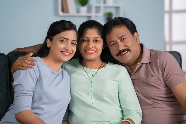 Happy smiling indian middle aged parents with adult daughter by looking at camera while sitting on sofa at home - concept of family relationship, togetherness and relaxation.