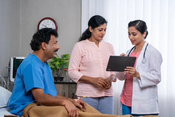 Indian doctor explaining to patients wife about medical report on digital tablet at hospital - concept of healthcare treatment, medicare service and Diagnosis Explanation