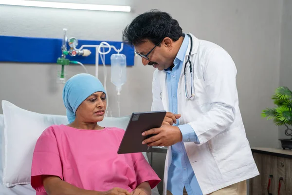 Indian doctor explaining health report from digital tablet to admitted cancer patient at hospital - concept of medicare support, health care guidance and treatment