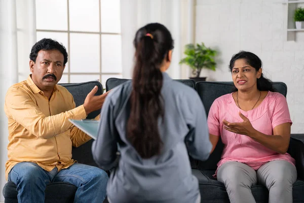 Shoulder shot of indian middle aged couple arguing in front of psychologist during counseling while sitting on sofa at home - concept of relationship problems, conversation and treatment