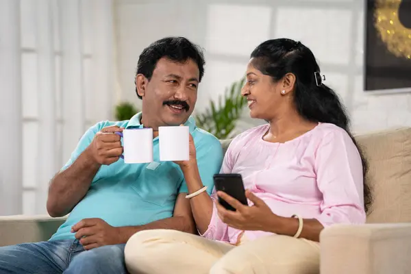 Happy indian caring husband giving coffee to his wife on sofa at home - concept of partner support, relationship bonding and Loving Gesture.