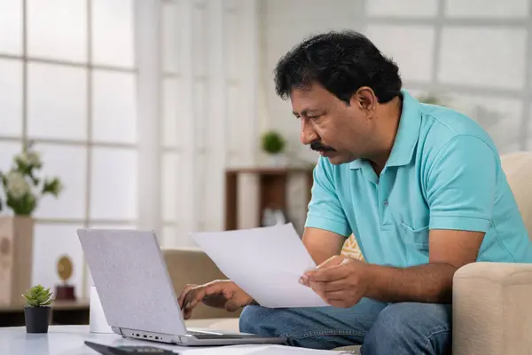 Indian middle aged man checking financial bills, insurence or mortgage documents on laptop while sitting on sofa at home - concept of Financial Planning, investment analysis and working on weekend