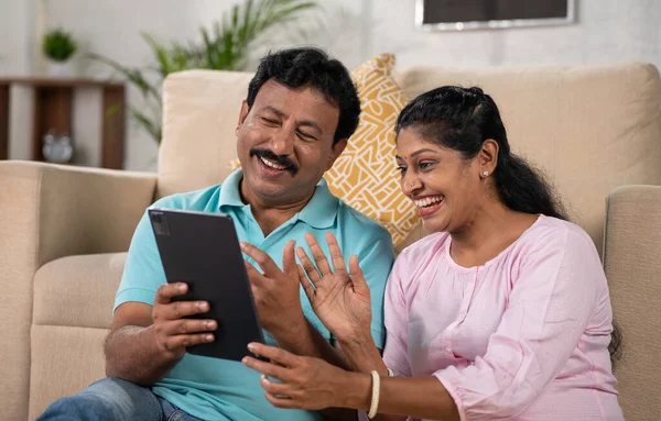 Happy smiling indian middle aged couple making video call on digital tablet while sitting on sofa at home - concept of family connection, togetherness and technology.