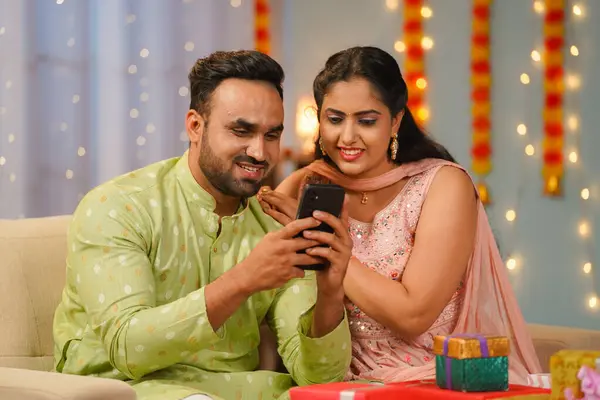 Happy young Indian couple using mobile phone together at home during diwali festival celebration at home - concept of social media sharing, festive wishes or greetings and Joyful Connection.