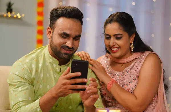 Happy young Indian couple using mobile phone together at home during diwali festival celebration in front of gifts- concept of social media sharing, festive wishes or greetings and Joyful Connection.