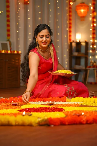 vertical shot of happy young Indian woman preparing diwali flower rangoli on floor for pooja or ritual at home - concept of festive celebration, special occasion and traditional culture or wear.