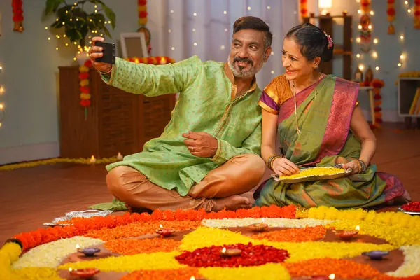 elderly senior couple taking selfie during diwali festive preparation at home in front of flower rangoli - concept of social media sharing, family bonding and traditional attire