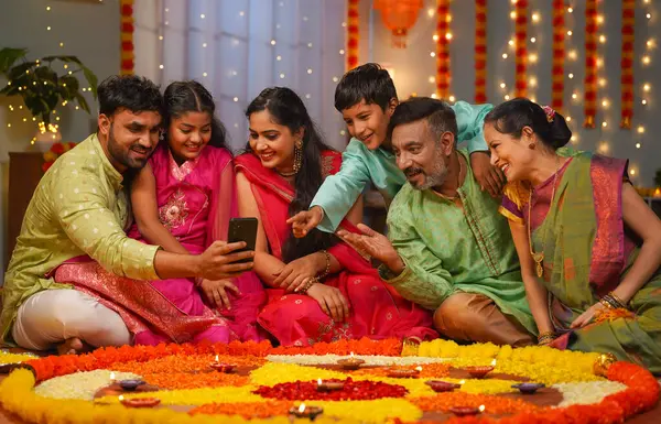 Group of happy indian joint family seeing mobile phone during family diwali festival celebration front of flower rangoli - concept of emotional reunions, Relationship bonding and social media sharing.