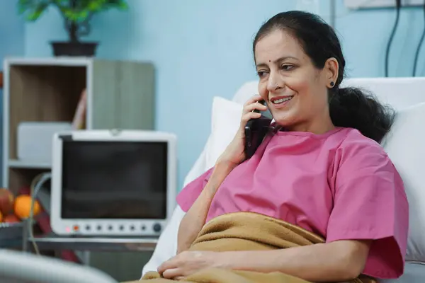 Happy Indian senior recovered patient talking on mobile phone call on hospital bed - concept of successful treatment, positive emotion and family interaction.