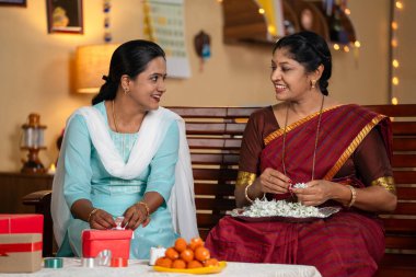 Joyful indian mother with daughter preparing of diwali festival celebration by packing gifts and stringing lowers at home - concept of traditional culture, relationship bonding and togetherness. clipart