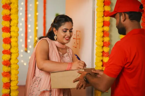 Happy girl receiving delivery box from delivery boy by signing on papers during Diwali festival day - concept of festive ecommerce order, fast delivery service and online shopping.