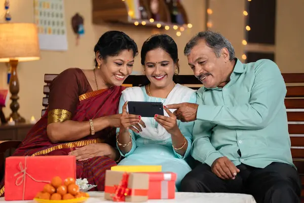 Joyful happy Indian mother and father watching mobile phone with daughter at home during diwali festival celebration - concept of cyberspace, social media sharing and online app streaming.