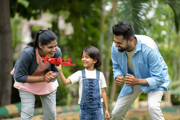 Tracking shot of happy indian kid playing airplane toy by running with couple at park - concept of childhood dreams, family support and freedom.