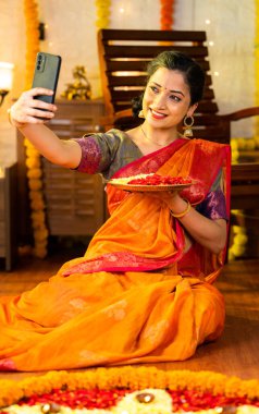 Çiçek tabağı tutarak cep telefonuyla selfie çeken mutlu Hintli kızın dikey çekimi - festival kutlaması, sosyal medya ve teknoloji konsepti.