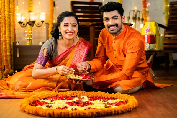 Happy smiling indian couple in traditional ethnic wear decorating rangoli with flowers on floor by looking camera at home - concept of family togetherness, religious and festival preparation