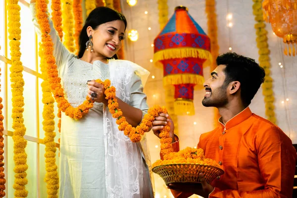 Wide shot of young indian man helping his wife for decorating flowers during Diwali festival celebration by talking each other at home - concept of togetherness, traditional culture and boding.