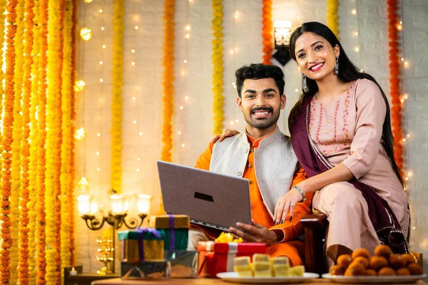 Happy smiling indian young couple using laptop by looking at camera while sitting on sofa at home - concept of festival offers, relationship and diwali celebration.