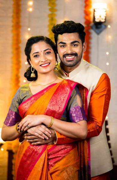 Happy indian couple in traditional ethnic clothing hugging by looking at camera at diwali festival celebration at home - concept of relationship, bonding and family time