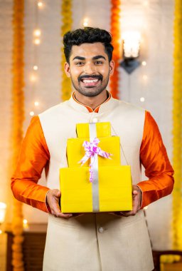 Mutlu, gülümseyen genç Hintli adamın evde Diwali kutlamaları sırasında kameraya bakarak hediye kutusu göstermesinin dikey çekimi - festival satışları, geleneksel ve alışveriş konsepti.