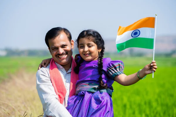 Happy Indian village father carrying daughter with indian flag by walking near green farmland during independence day - concept of democracy, patriotism and relationship bonding