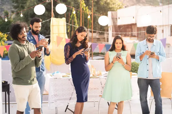 Group of young people or friends buys on mobile phones during birthday party meet or gathering on terrace - concept of cyberspace, smartphone or technology addiction and cyberspace.