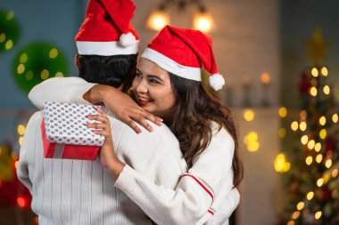 Mutlu bir kadının Noel Baba şapkasıyla evde kocalarına Noel hediyesi olarak sarıldığı bir omuz fotoğrafı. Romantik ilişki konsepti, ilgi arkadaşı ve şenlikli hediye paylaşımı.