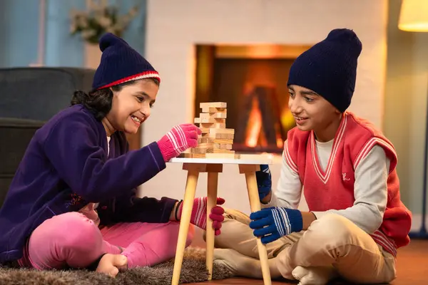 Indian siblings kids in winter wear busy playing block stacking game at home during winter holidays at home - concept of childhood concentration, entertainment and relationship bonding.