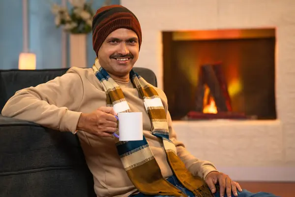 Relaxed portrait Indian man in winter wear drinking tea or coffee near warm fireplace - concept of weekend holidays, recreation and mindfulness.