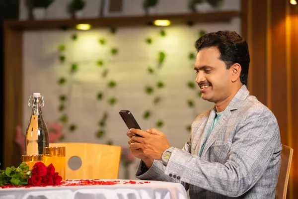 Indian happy middle aged man in suit using mobile phone during candle light dinner plan - concept of app conversation, date night and social media sharing.