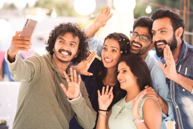 Akşamki parti sırasında terasta toplanan bir grup arkadaş görüntülü konuşma ya da sohbet - akıllı telefon iletişimi, sanal buluşma ve teknoloji kavramı.